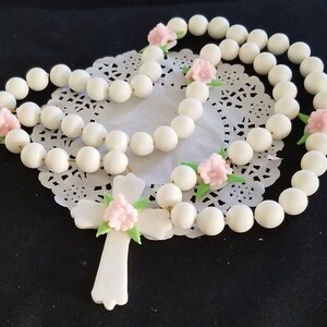 May include: A white rosary featuring large, spherical beads and a cross pendant. Pink floral accents and green leaves are placed along the rosary. The rosary is presented on a white, lace doily.