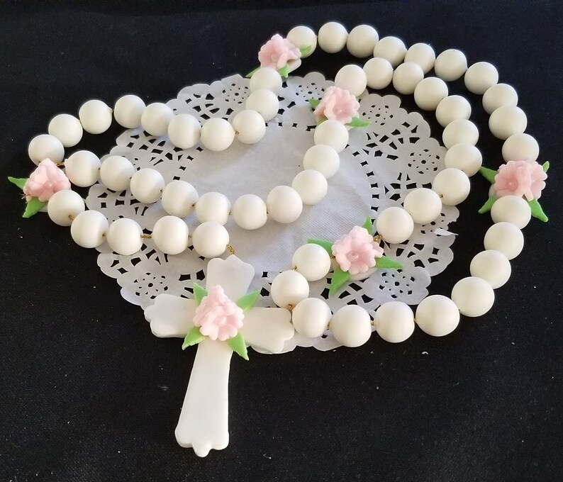 May include: A white rosary featuring large, spherical beads and a cross pendant. Pink floral accents and green leaves are placed along the rosary. The rosary is presented on a white, lace doily.