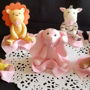 May include: Six baby animal figurines, each with a pink pacifier, sitting on a pink blanket. The animals are a lion, zebra, elephant, giraffe, monkey, and a second lion. The figurines are made of fondant and are perfect for a baby shower or birthday party.
