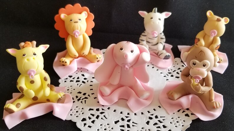 May include: Six baby animal figurines, each with a pink pacifier, sitting on a pink blanket. The animals are a lion, zebra, elephant, giraffe, monkey, and a second lion. The figurines are made of fondant and are perfect for a baby shower or birthday party.