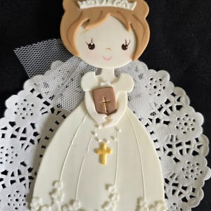 May include: A decorative figurine of a girl in a white dress, holding a book and a gold cross. The figurine has brown hair and a crown. It is set on a white lace doily, perfect for a religious celebration.