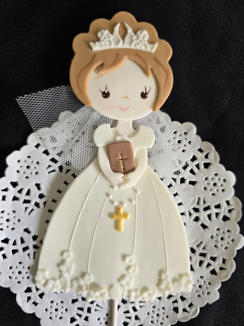 May include: A decorative figurine of a girl in a white dress, holding a book and a gold cross. The figurine has brown hair and a crown. It is set on a white lace doily, perfect for a religious celebration.