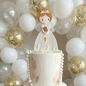 May include: A white communion cake featuring a fondant girl figure in a dress, holding a book and wearing a crown. The cake has pink and green decorations and a pearl border. White and gold balloons provide a celebratory background.