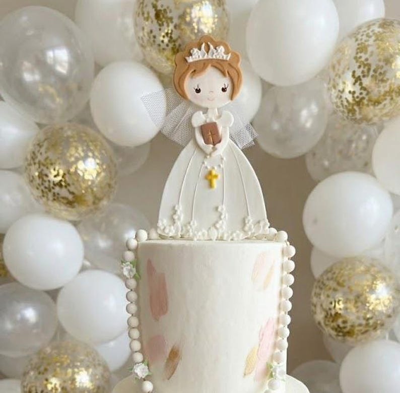 May include: A white communion cake featuring a fondant girl figure in a dress, holding a book and wearing a crown. The cake has pink and green decorations and a pearl border. White and gold balloons provide a celebratory background.