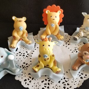 May include: Set of six baby animal cake toppers, each animal is sitting on a blue blanket. The animals include a lion, giraffe, zebra, monkey, elephant, and bear. Each animal is holding a blue pacifier.