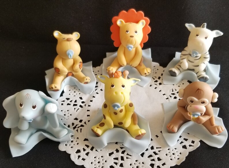 May include: Set of six baby animal cake toppers, each animal is sitting on a blue blanket. The animals include a lion, giraffe, zebra, monkey, elephant, and bear. Each animal is holding a blue pacifier.