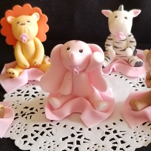 May include: Six baby animal figurines made of fondant, each wearing a pink blanket and holding a pink pacifier. The animals are a lion, zebra, elephant, giraffe, monkey, and a bear.
