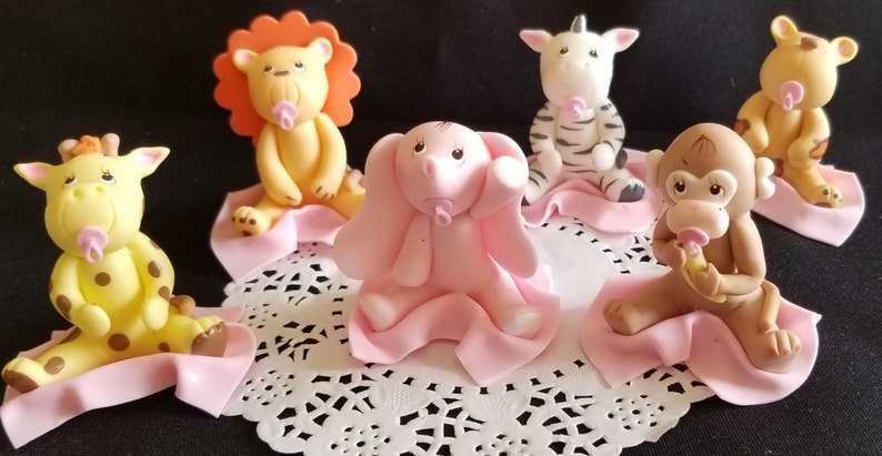May include: Six baby animal figurines made of fondant, each wearing a pink blanket and holding a pink pacifier. The animals are a lion, zebra, elephant, giraffe, monkey, and a bear.