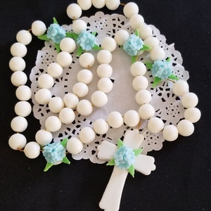 May include: A white rosary featuring large, spherical beads and a cross pendant. The rosary is decorated with light blue flower accents and green leaves. The beads are strung on gold-coloured wire.