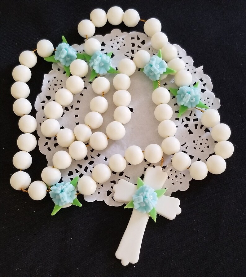 May include: A white rosary featuring large, spherical beads and a cross pendant. The rosary is decorated with light blue flower accents and green leaves. The beads are strung on gold-coloured wire.