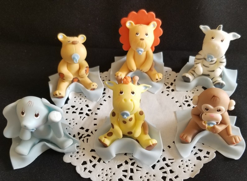 May include: Set of six baby animal cake toppers, each animal is sitting on a blue blanket. The animals include a lion, giraffe, zebra, monkey, elephant, and bear. Each animal is holding a blue pacifier.
