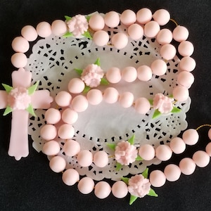 May include: A light pink rosary with a cross pendant and floral accents. The beads are round and connected by gold-coloured string. The cross and floral details are also light pink with green leaves. The rosary is displayed on a white lace doily.