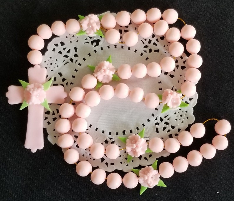 May include: A light pink rosary with a cross pendant and floral accents. The beads are round and connected by gold-coloured string. The cross and floral details are also light pink with green leaves. The rosary is displayed on a white lace doily.