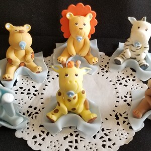 May include: Six adorable baby animal figurines, each sitting on a blue blanket. The animals include a lion, giraffe, zebra, monkey, elephant, and a bear. Each figurine has a pacifier in its mouth.