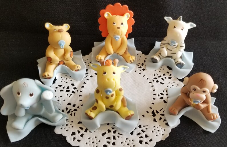 May include: Six adorable baby animal figurines, each sitting on a blue blanket. The animals include a lion, giraffe, zebra, monkey, elephant, and a bear. Each figurine has a pacifier in its mouth.