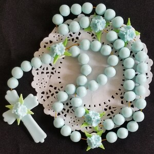May include: A pale blue rosary featuring round beads and a cross pendant. The rosary is decorated with light blue flowers and green leaves. The beads are strung on a gold-coloured string. The rosary is displayed on a white, lacy doily.
