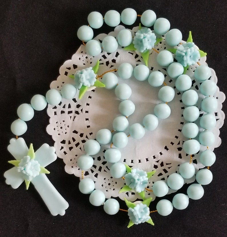 May include: A pale blue rosary featuring round beads and a cross pendant. The rosary is decorated with light blue flowers and green leaves. The beads are strung on a gold-coloured string. The rosary is displayed on a white, lacy doily.