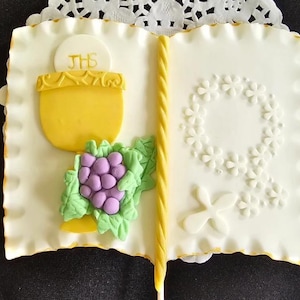 May include: A white, book-shaped cake decorated with a yellow chalice, purple grapes, and a floral wreath. The cake has a yellow border and a yellow candle spine. The chalice is inscribed with the letters "JHS".