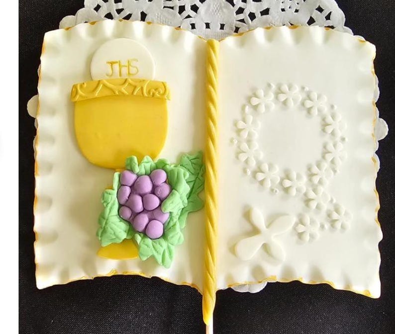 May include: A white, book-shaped cake decorated with a yellow chalice, purple grapes, and a floral wreath. The cake has a yellow border and a yellow candle spine. The chalice is inscribed with the letters "JHS".