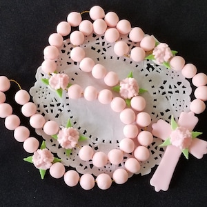 May include: A pink rosary featuring round beads and a cross, adorned with small pink flowers and green leaves. The rosary is presented on a white, lacy doily against a black backdrop. The beads are uniformly spaced.