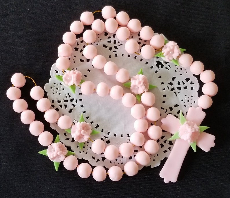 May include: A pink rosary featuring round beads and a cross, adorned with small pink flowers and green leaves. The rosary is presented on a white, lacy doily against a black backdrop. The beads are uniformly spaced.