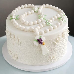 May include: A circular white cake decorated with a rosary made of white beads. The cake features a cross with purple grapes and a gold accent. The sides are adorned with white floral and lace-like patterns, creating an elegant design.