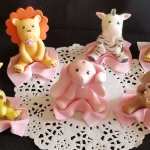 May include: Six baby animal figurines, each sitting on a pink blanket. The animals include a lion, zebra, elephant, giraffe, monkey, and a tiger. Each animal is holding a pink pacifier.
