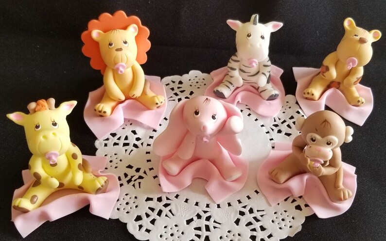 May include: Six baby animal figurines, each sitting on a pink blanket. The animals include a lion, zebra, elephant, giraffe, monkey, and a tiger. Each animal is holding a pink pacifier.