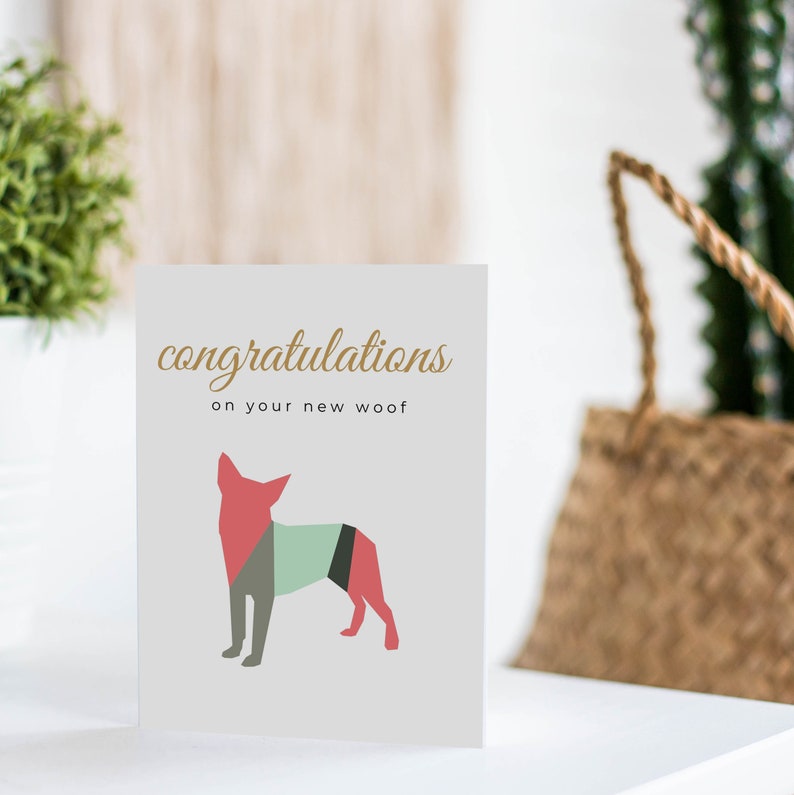 Puede incluir: Una tarjeta blanca con el texto "congratulations on your new woof" en letras doradas. Debajo del texto hay una ilustraci&oacute;n geom&eacute;trica de un perro en tonos rojos, verdes y grises.