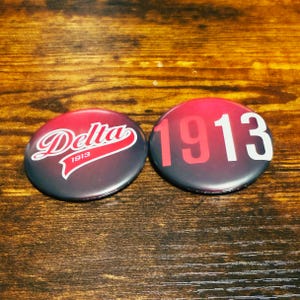 May include: Two round black and red buttons with white text. One button says "Delta 1913" in a white cursive font with a white banner underneath. The other button says "1913" in white text.