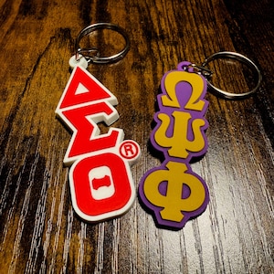 May include: Two keychains with Greek letters. The first keychain is red and white with the letters "ΔΣΘ" and a registered trademark symbol. The second keychain is purple and gold with the letters "ΩΨΦ".