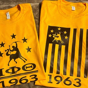 May include: Two yellow t-shirts with black designs. The left shirt features a black silhouette of a centaur riding a horse with a star and the text "IΦΟ 1963". The right shirt features a black silhouette of a centaur riding a horse with a star and a black and yellow American flag with the text "1963".