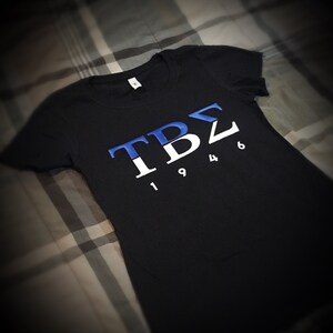 May include: Black t-shirt with a white and blue Greek letter design that says "TBZ 1946".