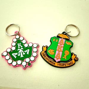 May include: Two keychains with silver rings. One is a pink and white ivy leaf shape with green and white lettering. The other is a green shield shape with pink and orange lettering and a gold border. Both have a silver key ring.