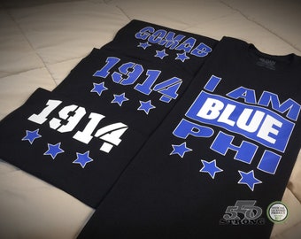 Puede incluir: Tres camisetas negras con letras azules y blancas. Las camisetas muestran el texto "1914", "I AM BLUE PHI" y "COMAB".