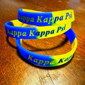 May include: Three flexible silicone wristbands featuring a vibrant blue and yellow swirl pattern. Each band is embossed with the text Kappa Kappa Psi in a bold, clear font. The bracelets are stacked together, showcasing the contrasting colors.