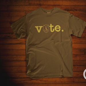 May include: A brown t-shirt with the word "vote" printed in yellow on the front. The word "vote" includes a graphic of a buffalo head with a shield.