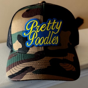 May include: A black and green camouflage trucker hat with a blue and yellow embroidered patch that says "Pretty Poodles".