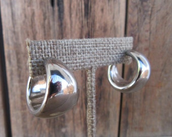May include: A pair of polished silver-tone hoop earrings with a thick, rounded, and chunky profile. The earrings feature a smooth, reflective metallic surface that catches the light, showcasing a wide, curved design that tapers slightly toward the ends.