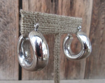 May include: A pair of silver hoop earrings with a smooth, rounded design. The earrings are displayed on a burlap stand.
