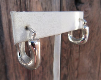 May include: A pair of polished silver hoop earrings. The earrings have a rounded, rectangular shape and are displayed on a white earring stand against a wooden background. The earrings are reflective and have a smooth surface.