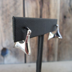 Puede incluir: Un par de pendientes de plata con forma triangular. Los pendientes se muestran en un soporte negro.