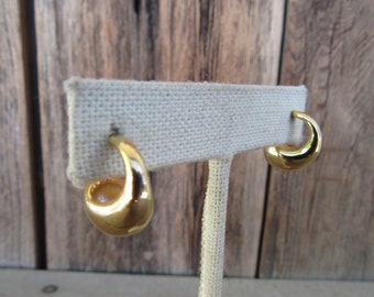 May include: A pair of gold-colored hoop earrings. The earrings have a smooth, polished surface and a rounded, crescent shape. They are displayed on a neutral-colored earring stand against a wooden background. The earrings are a classic jewelry item.