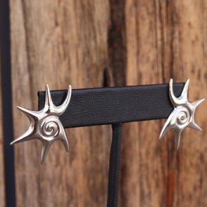 以下が含まれることがあります： 星形のデザインと中央に渦巻き模様が施された銀製のイヤリングのペア。イヤリングは黒いディスプレイスタンドに置かれています。