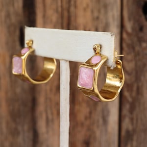 May include: A pair of gold hoop earrings with pink enamel accents. The earrings are displayed on a white stand.