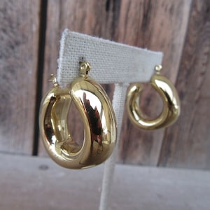 May include: A pair of gold hoop earrings with a thick, rounded design. The earrings are displayed on a white fabric stand.