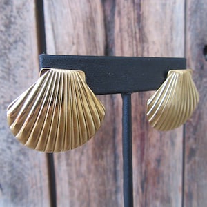 May include: A pair of gold-tone shell-shaped earrings. The earrings are on a black display stand.