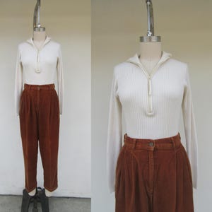 May include: A white ribbed sweater with a zipper neckline and long sleeves. The sweater is paired with brown corduroy pants with pleats.