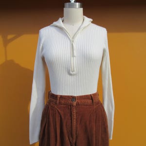 May include: A white ribbed sweater with a zipper and long sleeves, paired with brown corduroy pants.
