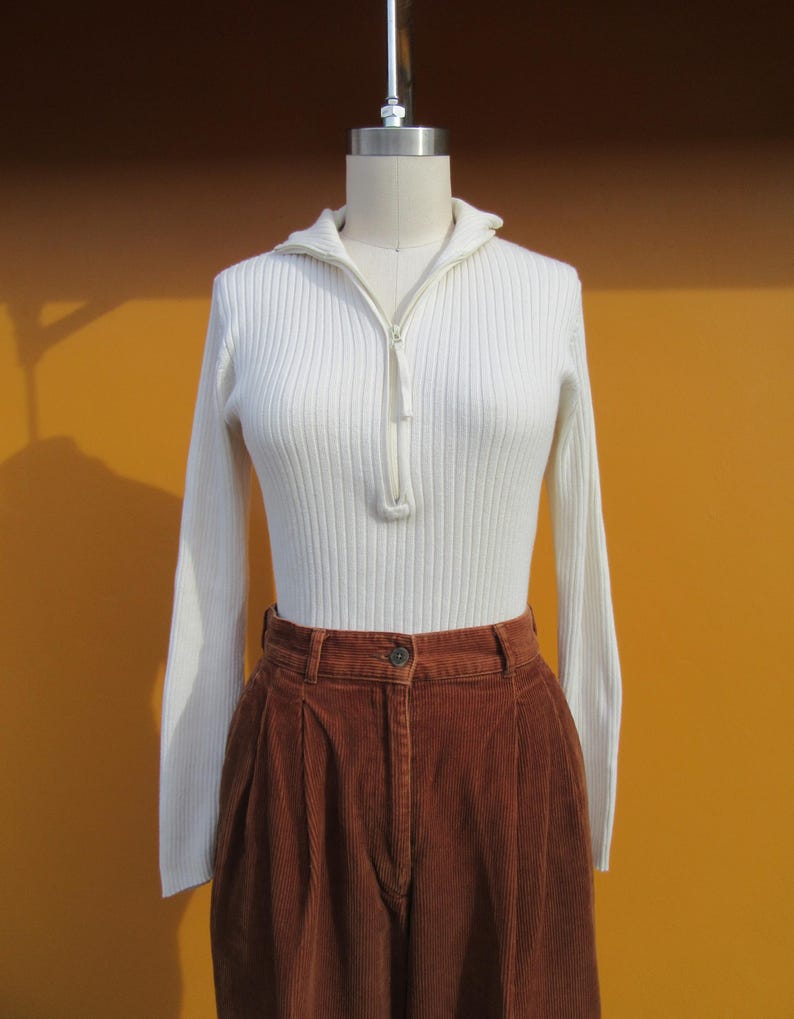May include: A white ribbed sweater with a zipper and long sleeves, paired with brown corduroy pants.